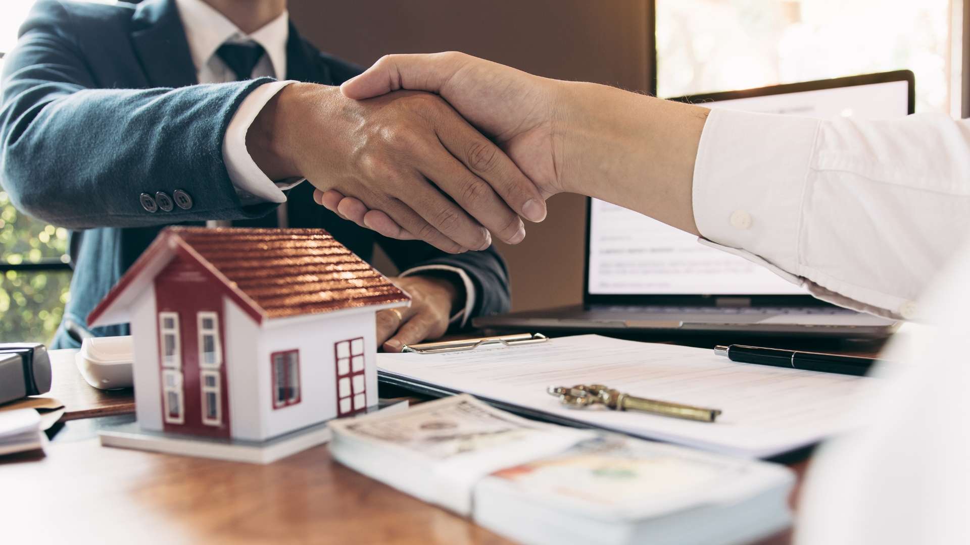 Two business professionals shaking hands over a real estate contract, symbolizing a property investment agreement, with a model house and legal documents on the table.