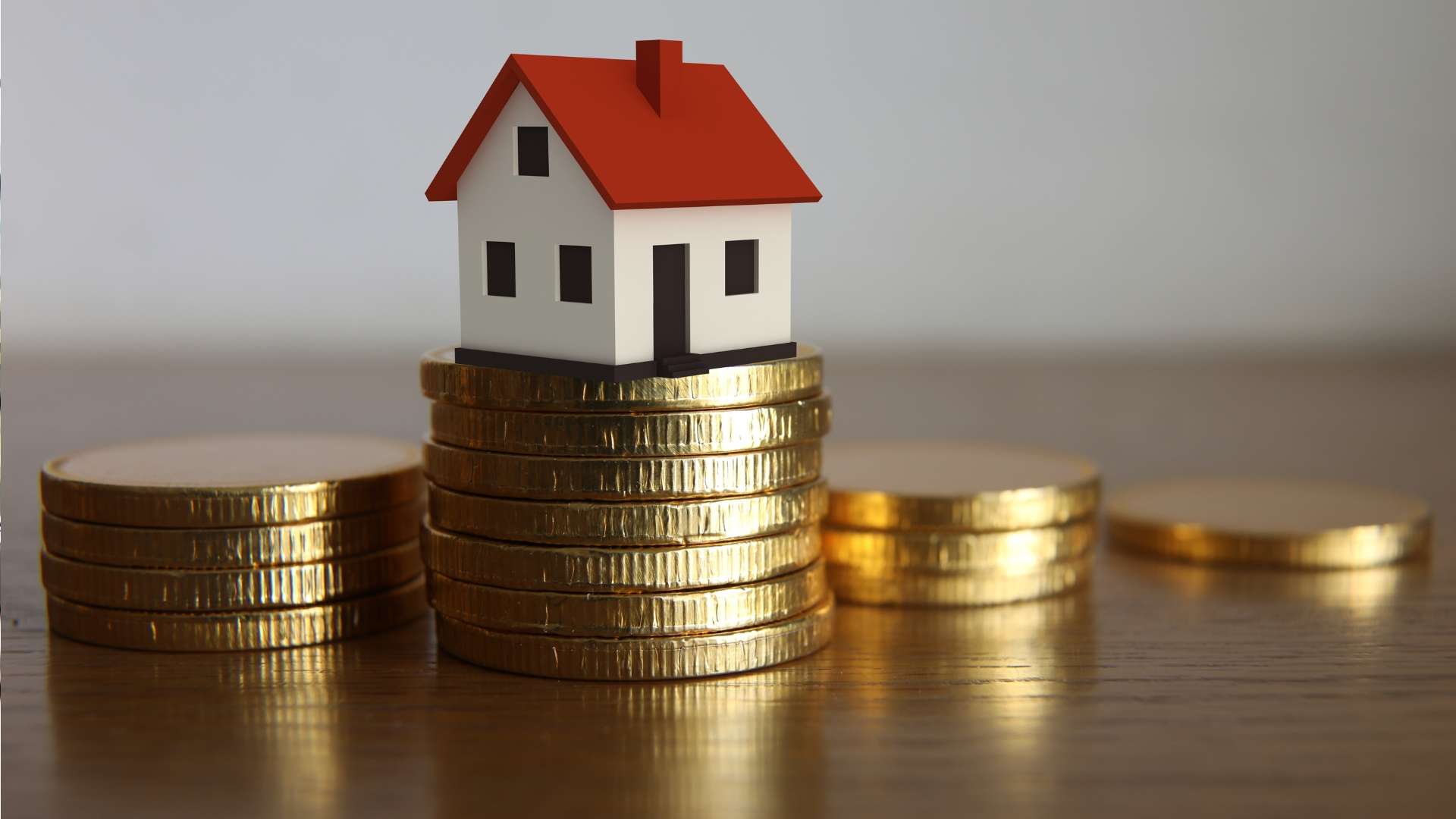 Small model house with a red roof placed on top of stacked gold coins, symbolizing real estate investment and property value.