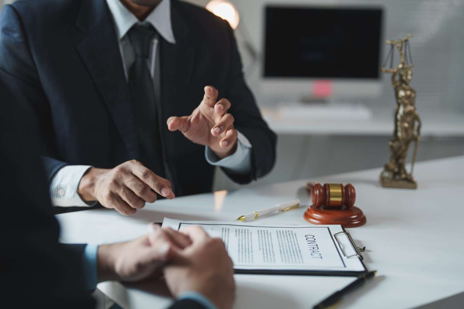 An LA trademark attorney explaining contract details to a client across a desk with a gavel, contract clipboard, and Lady Justice statue, representing the expert legal guidance Carbon Law Group provides for trademarking a brand name in California.